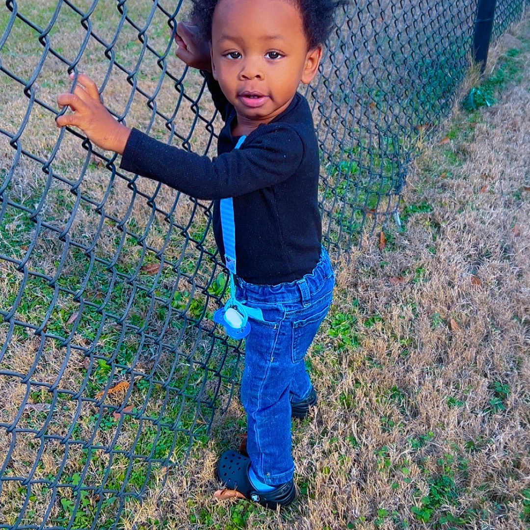 Mosiah is registered to the contest to win money with this photo: afro_hair, black_shirt, blue_jeans, casual_clothing, chain_link_fence, child, crocs, curious, daylight, fence, field, grass, holding, nature, outdoor, pacifier, person, standing, toddler, young_child