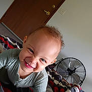 Evelyn joined the competition — help win amazing prizes! child, toddler, smiling, face, curly_hair, bedroom, blanket, fan, door, wall, light_switch, clothing, person, indoor, happy, playful, young_child, expression, home, casual