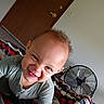 child, toddler, smiling, face, curly_hair, bedroom, blanket, fan, door, wall, light_switch, clothing, person, indoor, happy, playful, young_child, expression, home, casual
