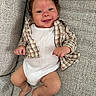 baby, smiling, infant, couch, plaid_shirt, white_onesie, curly_hair, happy, person, indoor, skin, face, legs, feet, hand, textile, fabric, relaxing, child, portrait