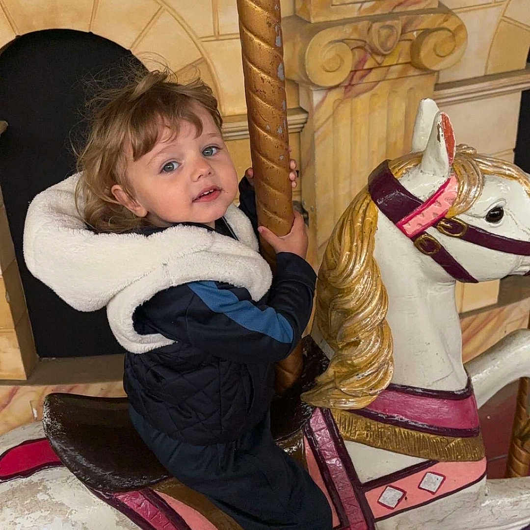 Tino participe au concours pour gagner de l'argent avec cette photo : amusement_park, carousel, child, classic, curly_hair, decor, entertainment, fun, golden_pole, horse, indoor, jacket, person, playful, portrait, ride, smiling, sneakers, toy, vintage