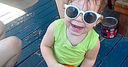 Ares is registered to the contest to win money with this photo: toddler, child, sunglasses, smiling, orange_ball, green_tank_top, blue_shorts, blue_footwear, porch, wooden_floor, adult, summer, outdoor, happy, person, casual, playful, sunny, family, fun