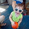 toddler, child, sunglasses, smiling, orange_ball, green_tank_top, blue_shorts, blue_footwear, porch, wooden_floor, adult, summer, outdoor, happy, person, casual, playful, sunny, family, fun