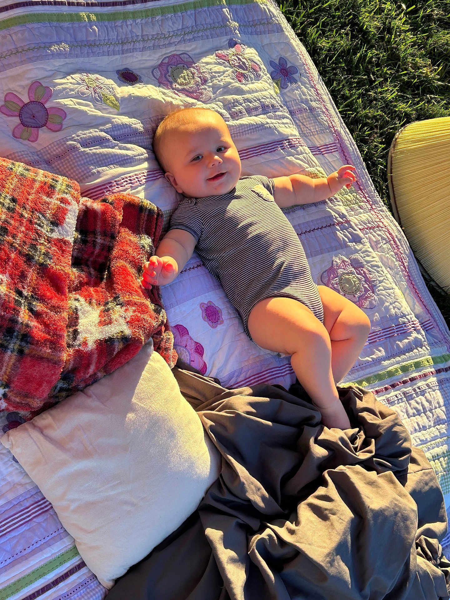 Bruce is registered to the contest to win money with this photo: baby, infant, quilt, blanket, pillow, outdoor, grass, sunlight, smiling, clothing, striped_onesie, relaxing, child, cute, happy, lying_down, soft_texture, warm_light, resting, nature