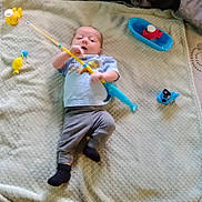 Xavier is registered to the contest to win money with this photo: baby, child, toy, fishing_rod, blanket, blue_shirt, grey_pants, black_socks, plastic_fish, blue_basin, yellow_fish_toy, indoor, playtime, infant, lying_down, curious, soft_surface, childhood, cute, home