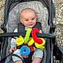 baby, child, stroller, plush_toy, colorful_toy, outdoor, cobblestone, infant, clothing, seat, straps, cute, person, portrait, happy, smile, head, hand, feet, fabric