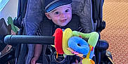 Jordan is registered to the contest to win money with this photo: baby, stroller, cap, toy, plush_toy, smile, infant, child, cushion, clothing, sock, seatbelt, indoor, carpet, pattern, hand, face, person, cute, happy