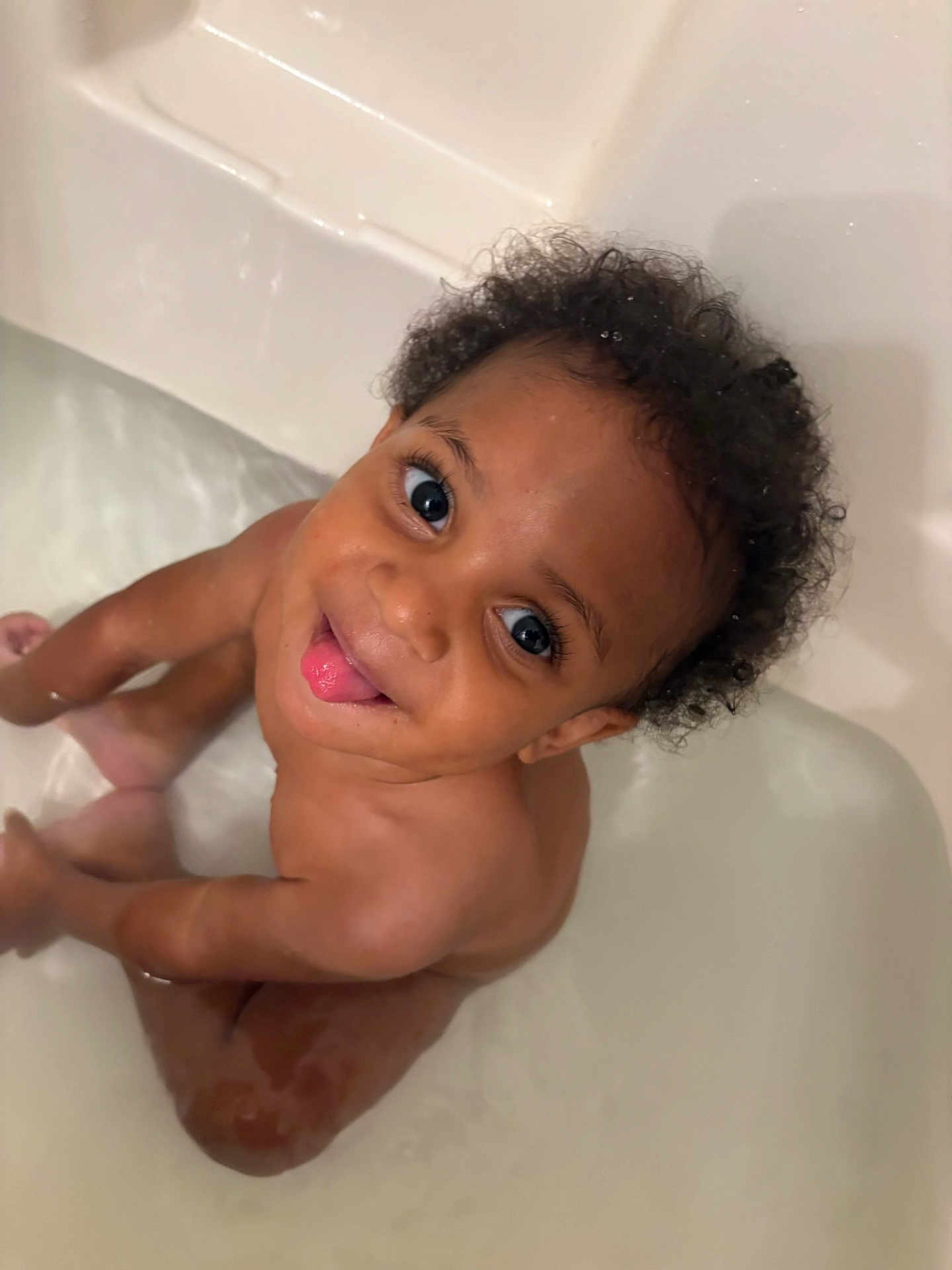 Legend is registered to the contest to win money with this photo: toddler, child, bath, water, curly_hair, tongue_out, playful, smiling, skin, sitting, bathtub, cute, happy, indoors, person, young_child, fun, bath_time, expression, splash