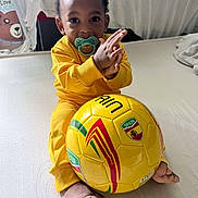 Taylor a rejoint le concours — aidez-le/la à gagner de superbes lots ! toddler, child, baby, pacifier, yellow_clothing, soccer_ball, sports, indoor, bed, footwearless, clapping, curly_hair, smiling, cute, playful, person, toy, bright_colors, home, young_child