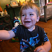 Rexton is registered to the contest to win money with this photo: toddler, child, smiling, happy, indoor, shirt, graphic_tshirt, floor, wooden_floor, furniture, speaker, toy, playpen, person, face, arm, hair, expression, home, room