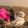 person, dog, couch, living_room, pet, white_dog, casual, indoor, relaxing, comfort, paw, collar, brown_couch, pink_shirt, camouflage_pants, table, bottle, medicine, head_touch, bonding