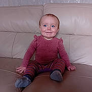 Alice is registered to the contest to win money with this photo: baby, child, toddler, blue_eyes, pink_shirt, striped_pants, socks, leather_couch, indoor, smiling, happy, seated, person, face, furniture, home, cute, young_child, casual_clothing, portrait