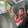 black_car_seat, bottle, car_interior, car_seat, child, curly_hair, daylight, headrest, infant, peaceful, red_clothing, resting, safety, seat, seatbelt, sleeping, toddler, toy, travel, window