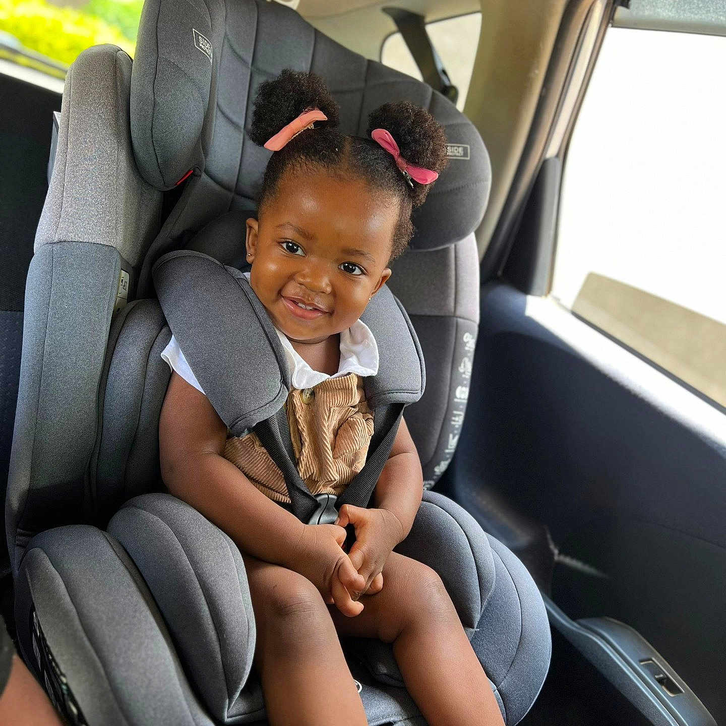 Zoey is registered to the contest to win money with this photo: baby, car_interior, car_seat, child, clothing, cute, daylight, hair_buns, happy, person, pink_ribbons, portrait, safety, seatbelt, shoes, sitting, smile, toddler, vehicle, window