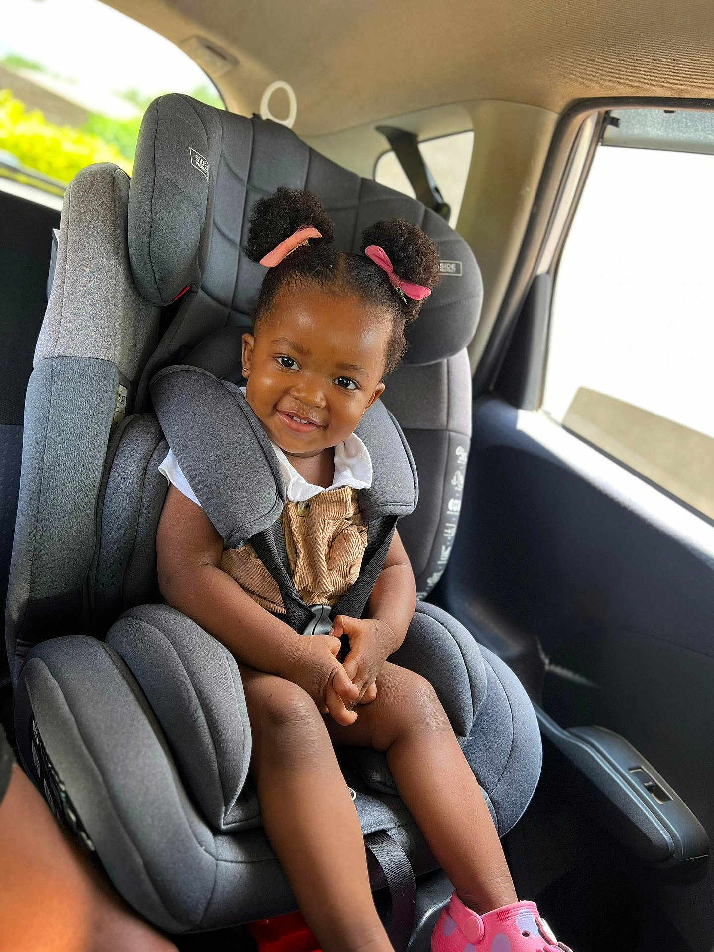 Zoey is registered to the contest to win money with this photo: child, toddler, car_seat, smile, hair_buns, pink_ribbons, clothing, shoes, car_interior, seatbelt, happy, person, portrait, baby, cute, sitting, safety, window, daylight, vehicle