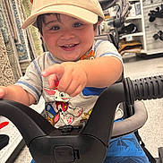 Arsenio is registered to the contest to win money with this photo: toddler, child, cap, smile, pointing, shopping_cart, store, clothing, shorts, toy, happy, face, person, indoor, retail, footwear, pants, handlebar, cute, boy