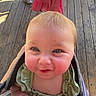 baby, blond_hair, bright_eyes, carrier, casual, child, curious, cute, daylight, face, green_clothing, hand, infant, outdoor, person, portrait, rosy_cheeks, small_hands, smiling, wooden_floor