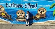 Jaxson is registered to the contest to win money with this photo: child, mural, otters, cartoon, painting, wall, sidewalk, outdoor, sunny, blue_sky, building, welcome_text, casual_clothing, shorts, sneakers, backpack, shadow, concrete, daytime, smiling