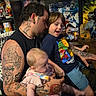 man, baby, child, tattoos, chair, collectibles, posters, indoor, family, smiling, casual_clothing, sitting, table, figurines, comic_books, colorful, fun, home, happy, boy