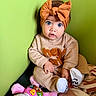 Kehlani is registered to the contest to win money with this photo: baby, child, headband, orange, bow, blue_eyes, beige_clothing, sitting, green_wall, plush_toy, pink, blanket, cute, indoors, portrait, soft_toys, expression, cozy, toddler, person