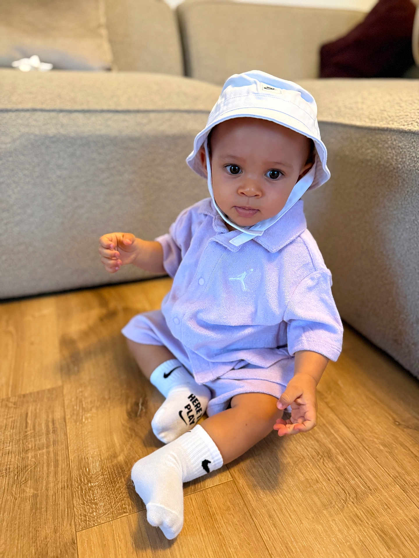 Ayden a rejoint le concours — aidez-le/la à gagner de superbes lots ! toddler, child, baby, sitting, floor, hat, bucket_hat, socks, white_socks, purple_outfit, shorts, shirt, indoor, wooden_floor, couch, furniture, curious, portrait, person, clothing