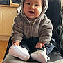 baby, child, stroller, hoodie, smiling, indoor, white_shoes, cute, face, person, seated, clothing, happy, infant, furniture, black_stroller, toy, footwear, babywear, warm_clothing