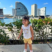 Valeria is registered to the contest to win money with this photo: child, toddler, girl, smile, waterfront, marina, boats, yacht, building, skyscraper, modern_architecture, greenery, hedge, sneakers, shorts, tank_top, sunny, outdoor, promenade, string_lights