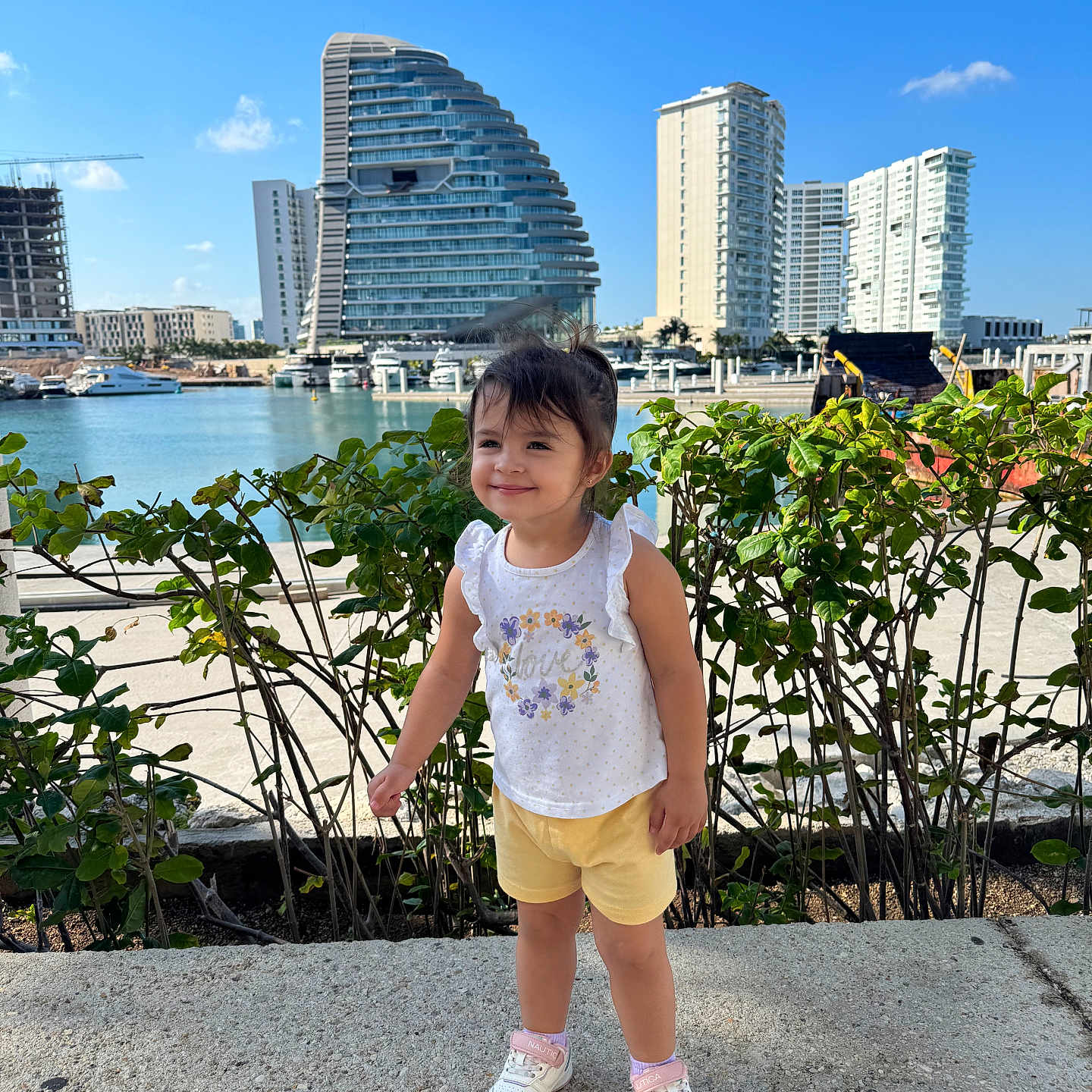 Valeria is registered to the contest to win money with this photo: boats, building, child, girl, greenery, hedge, marina, modern_architecture, outdoor, promenade, shorts, skyscraper, smile, sneakers, string_lights, sunny, tank_top, toddler, waterfront, yacht