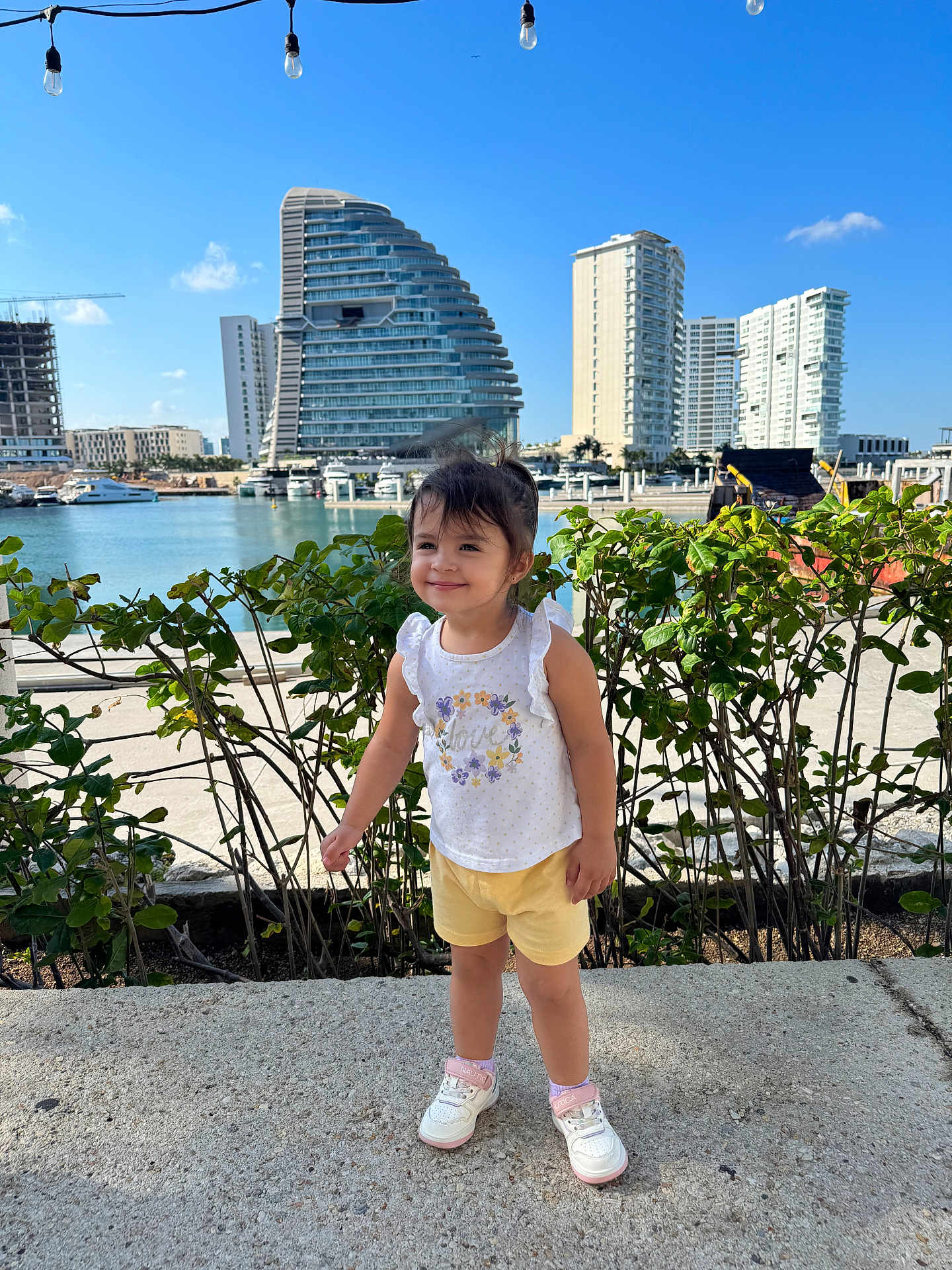 Valeria is registered to the contest to win money with this photo: child, toddler, girl, smile, waterfront, marina, boats, yacht, building, skyscraper, modern_architecture, greenery, hedge, sneakers, shorts, tank_top, sunny, outdoor, promenade, string_lights