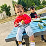 blue_shoes, building, casual_clothing, child, cucumber, curious, greenery, nature, outdoor, pavement, person, picnic_table, playful, siblings, sitting, snack, summer, toddler, trees, yellow_socks