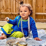 Aarush Jha joined the competition — help win amazing prizes! child, toddler, smiling, traditional_clothing, blue_tunic, green_pants, pearl_necklace, outdoor, quilt, spoon, cream, face_paint, decorative_bowls, candles, wooden_fence, grass, curly_hair, playful, sitting, portrait