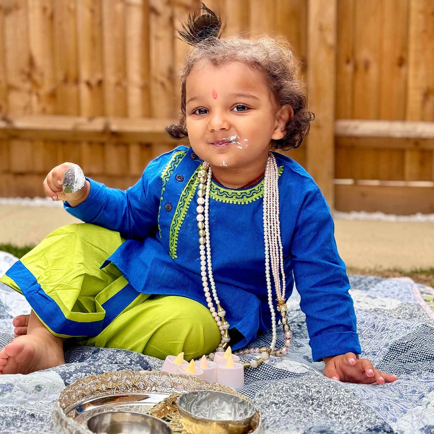 Aarush Jha joined the competition — help win amazing prizes! blue_tunic, candles, child, cream, curly_hair, decorative_bowls, face_paint, grass, green_pants, outdoor, pearl_necklace, playful, portrait, quilt, sitting, smiling, spoon, toddler, traditional_clothing, wooden_fence