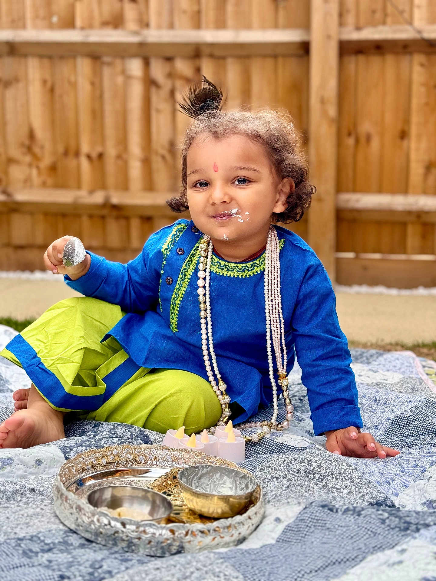 Aarush Jha joined the competition — help win amazing prizes! child, toddler, smiling, traditional_clothing, blue_tunic, green_pants, pearl_necklace, outdoor, quilt, spoon, cream, face_paint, decorative_bowls, candles, wooden_fence, grass, curly_hair, playful, sitting, portrait