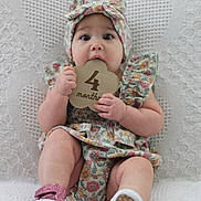 Charli is registered to the contest to win money with this photo: baby, infant, child, floral_outfit, headband, wooden_sign, 4_months, cute, sitting, blanket, texture, footwear, pink_shoes, playful, curious, portrait, indoors, hands, face, expression