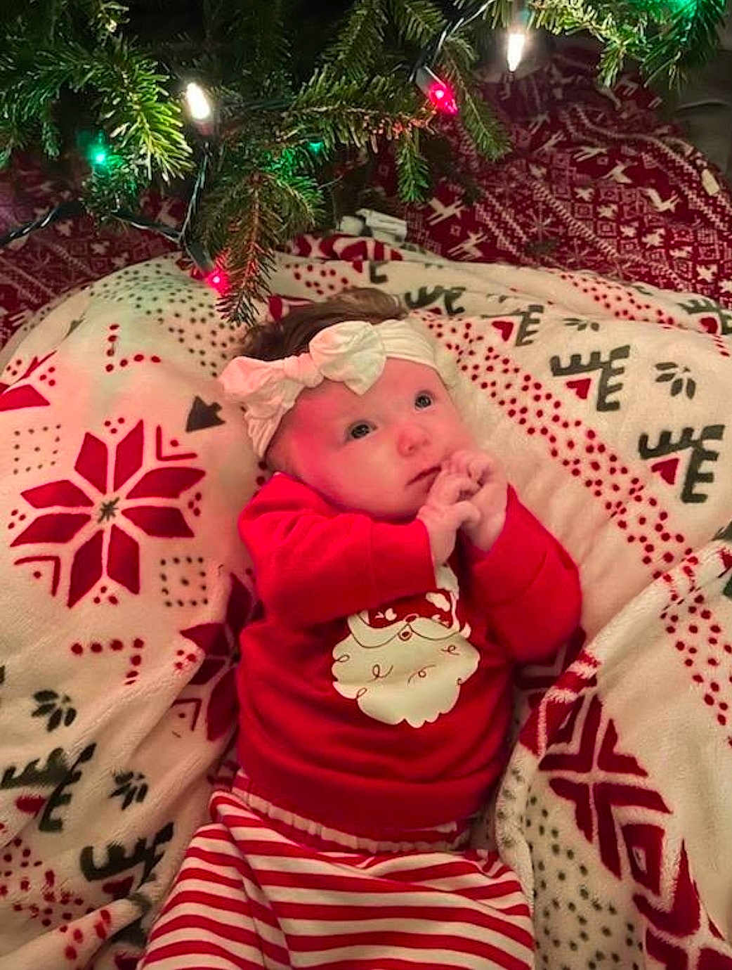 Emma is registered to the contest to win money with this photo: baby, infant, child, christmas, holiday, blanket, christmas_tree, string_lights, headband, red_clothing, santa_print, striped_pants, cozy, festive, portrait, lying_down, indoors, gaze, soft_texture, bedding