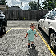 Victor is registered to the contest to win money with this photo: child, parking_lot, car, asphalt, shadow, cloudy_sky, fence, tree, clothing, shoes, shorts, polo_shirt, outdoor, daylight, sidewalk, vehicle, person, expression, sunlight, nature