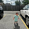 child, parking_lot, car, asphalt, shadow, cloudy_sky, fence, tree, clothing, shoes, shorts, polo_shirt, outdoor, daylight, sidewalk, vehicle, person, expression, sunlight, nature