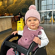 Rose is registered to the contest to win money with this photo: baby, child, toddler, pink_hat, knit_hat, pom_pom, fuzzy_outfit, baby_seat, high_chair, cafe, coffee_shop, mural, serpent_art, caution_sign, wet_floor_sign, smile, face, indoor, glass_door, floor