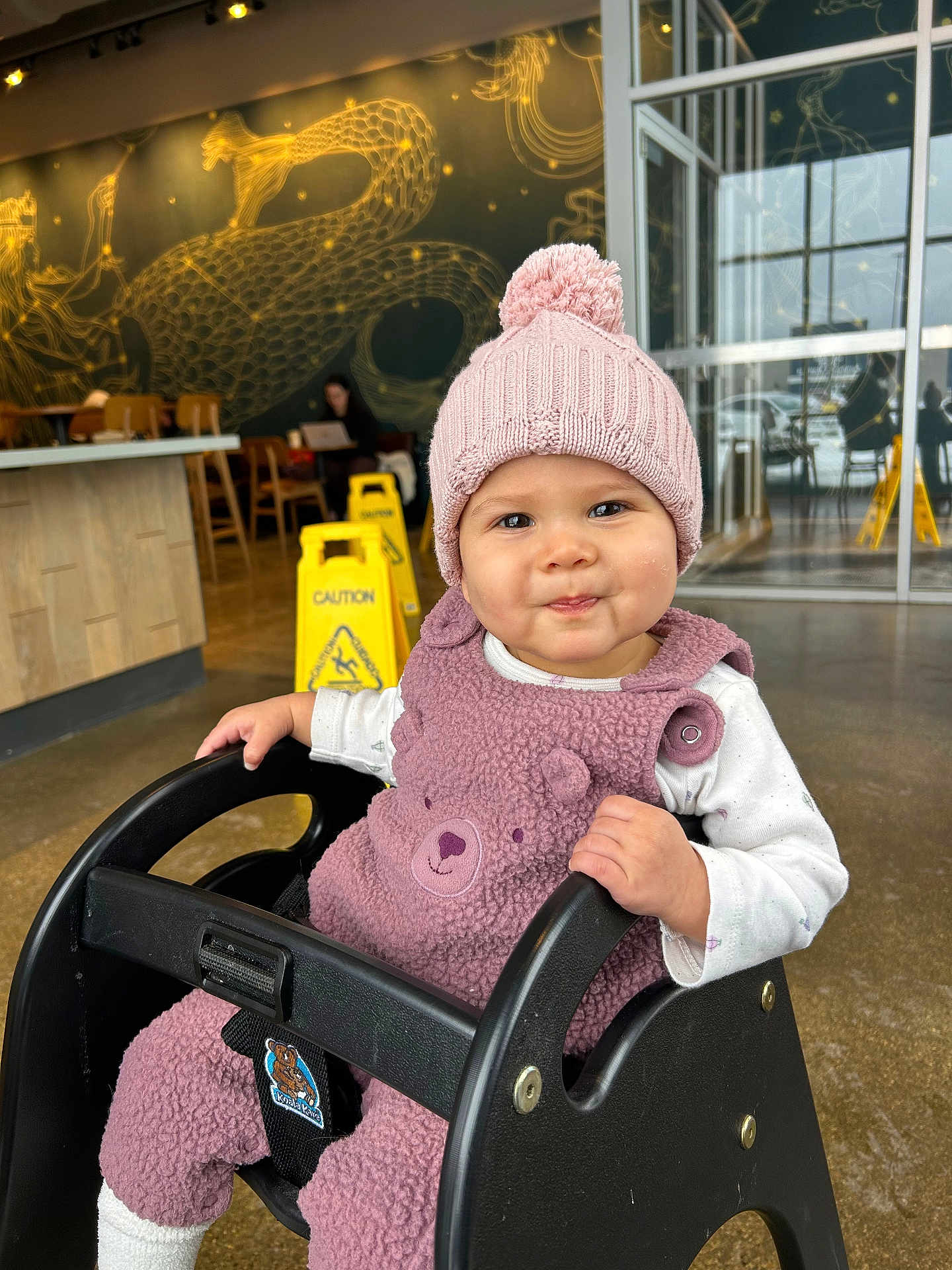 Rose is registered to the contest to win money with this photo: baby, child, toddler, pink_hat, knit_hat, pom_pom, fuzzy_outfit, baby_seat, high_chair, cafe, coffee_shop, mural, serpent_art, caution_sign, wet_floor_sign, smile, face, indoor, glass_door, floor