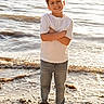 child, boy, beach, sand, water, ocean, barefoot, casual_clothing, white_shirt, jeans, smiling, standing, sunlight, outdoor, nature, waves, happy, summer, portrait, daytime