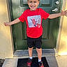 child, boy, red_tshirt, spiderman, black_shorts, sneakers, door, doormat, outdoor, smiling, happy, standing, short_hair, casual_clothing, sunlight, shadow, concrete, wall, playful, portrait
