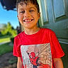 child, boy, smiling, red_tshirt, spiderman, outdoor, green_door, sunny, portrait, happy, short_hair, casual_clothing, daylight, background_blur, front_view, young, person, standing, nature, bright_colors