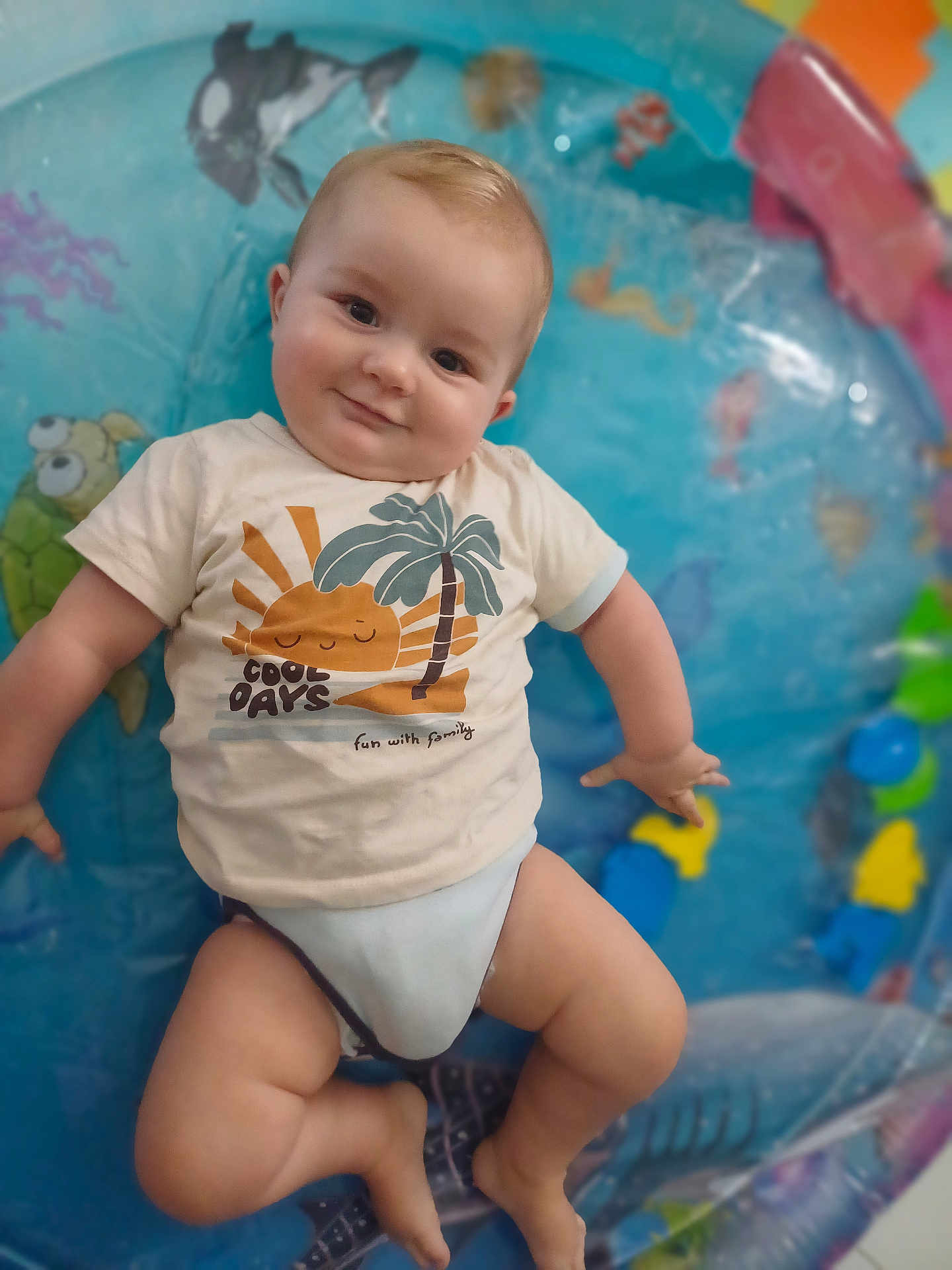 Léo a rejoint le concours — aidez-le/la à gagner de superbes lots ! baby, infant, tshirt, smile, chubby_cheeks, indoor, inflatable_pool, colorful, cute, playful, child, sea_creatures, turtle, fish, palm_tree, sun_graphic, relaxed, skin, legs, happy