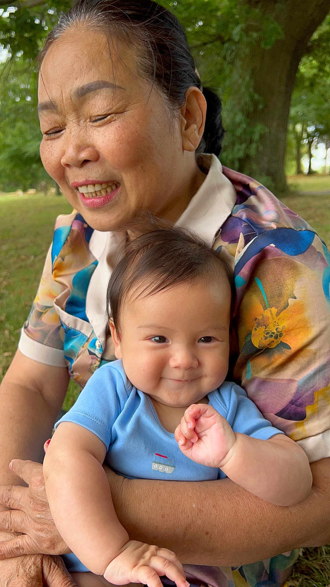 Alfred is registered to the contest to win money with this photo: elderly_woman, baby, smiling, outdoor, nature, greenery, trees, colorful_clothing, floral_pattern, embrace, happy, portrait, family, baby_clothes, person, skin, head, face, closeup, love