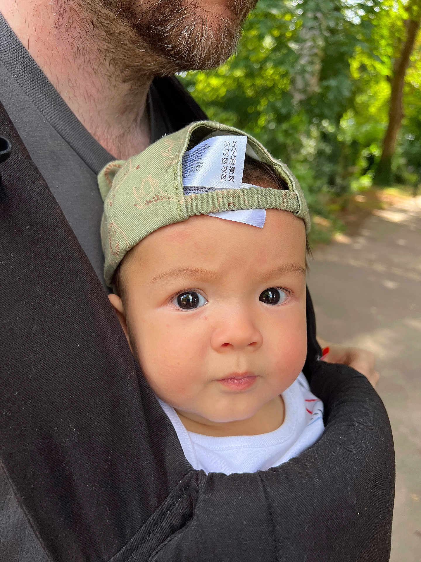 Alfie is registered to the contest to win money with this photo: baby, child, infant, cap, green_cap, baby_carrier, adult, beard, outdoor, forest, path, nature, portrait, closeup, face, curious, expression, skin, clothing, daylight