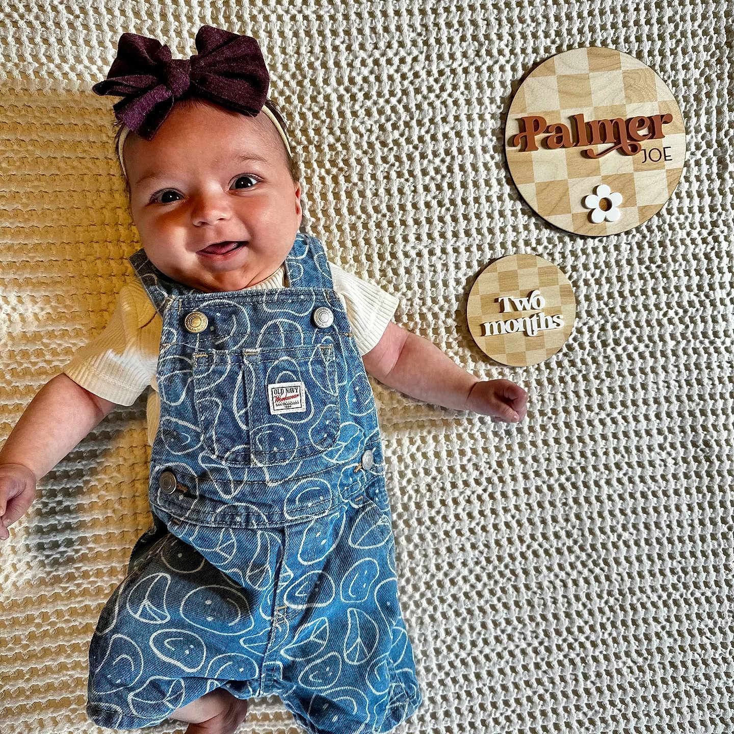 Palmer is registered to the contest to win money with this photo: baby, blanket, bow, child, cute, denim, happy, headband, infant, lying_down, milestone, overalls, pattern, person, plaque, portrait, smiling, text, white_shirt, wood