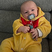 Jules participe au concours pour gagner de l'argent avec cette photo : baby, child, comfort, controller, couch, cute, grey_couch, home, indoor, infant, pacifier, person, plush_clothing, red_onesie_detail, relaxed, seated, toy, video_game_controller, yellow_onesie, young_child