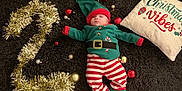 Jules a rejoint le concours — aidez-le/la à gagner de superbes lots ! baby, carpet, celebration, christmas, cute, elf_costume, festive, gold_balls, gold_tinsel, green_hat, holiday, holiday_decor, indoors, infant, lying_down, number_two, ornaments, pillow, red_balls, striped_pants