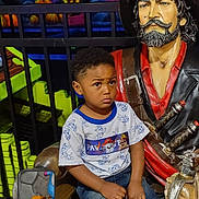 Jacoby is registered to the contest to win money with this photo: child, boy, pirate, statue, amusement_park, colorful, neon, indoor, reflection, phone, casual_clothing, blue_shoes, jeans, tshirt, seated, expression, playful_background, decor, portrait, young