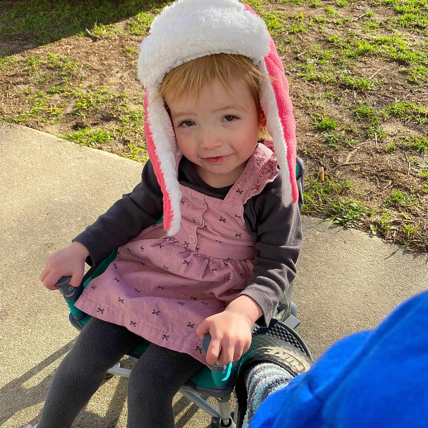 Delilah is registered to the contest to win money with this photo: car_tire, casual, chair, child, concrete, footwear, grass, hand, hat, jacket, legs, outdoor, person, pink_dress, playful, seat, smile, sunlight, toddler, toy